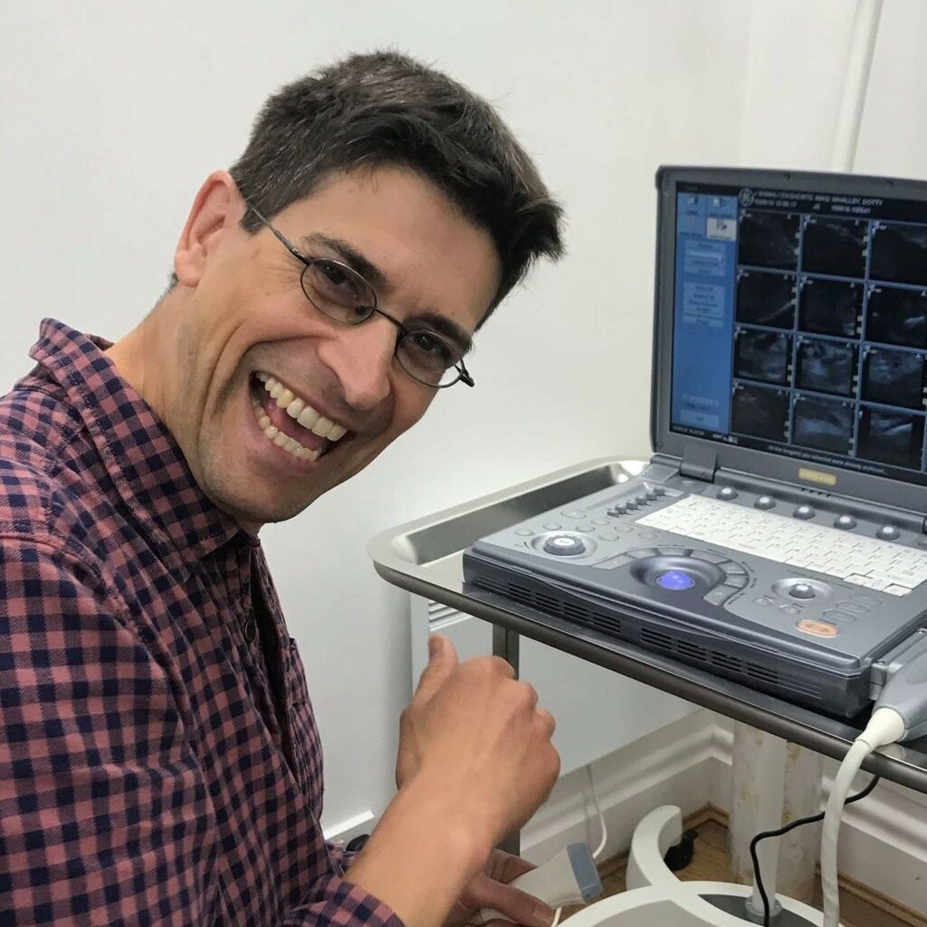 A bespectacled individual smiles while seated beside a medical imaging device, with a laptop showing scan results, surrounded by three specialists at a veterinary referral centre.
