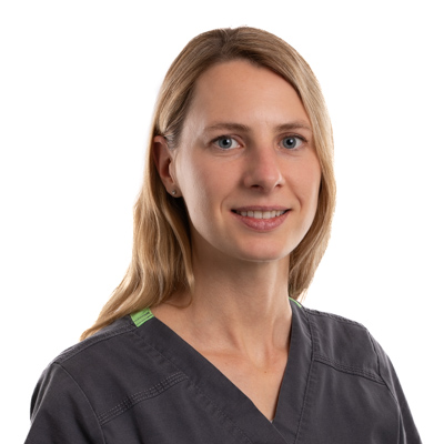 A woman with long blonde hair, dressed in dark medical scrubs, stands before a plain white background. She faces the camera with a gentle smile.