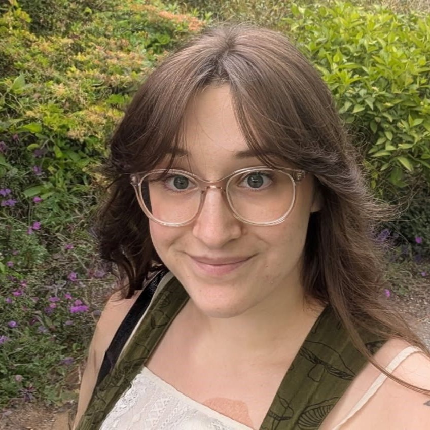 Emily Kennedy, wearing glasses and with long brown hair, smiles at the camera while standing outside surrounded by green foliage and purple flowers.