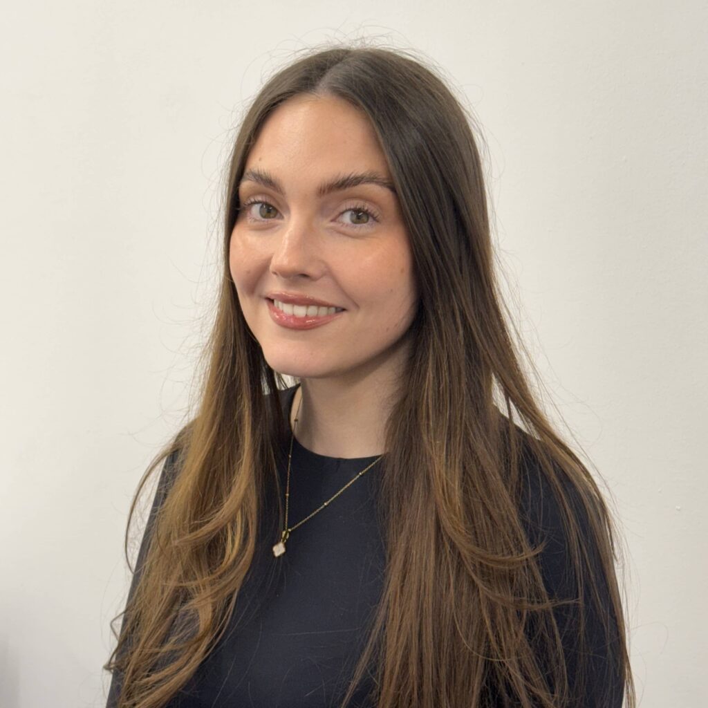 A woman with long brown hair, dressed in a black top and a necklace, smiles at the camera in front of a plain, light-coloured background.